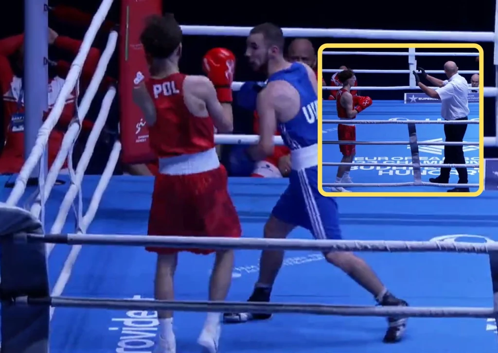 Dwóch bokserów w czerwonym i niebieskim stroju walczy na ringu bokserskim, zawodnik w czerwonym stroju pochodzi z Polski, a w niebieskim z Ukrainy. W tle widać sędziego oraz powtórkę w rogu obrazu, gdzie sędzia odlicza czas, a jeden z zawodników przygo...