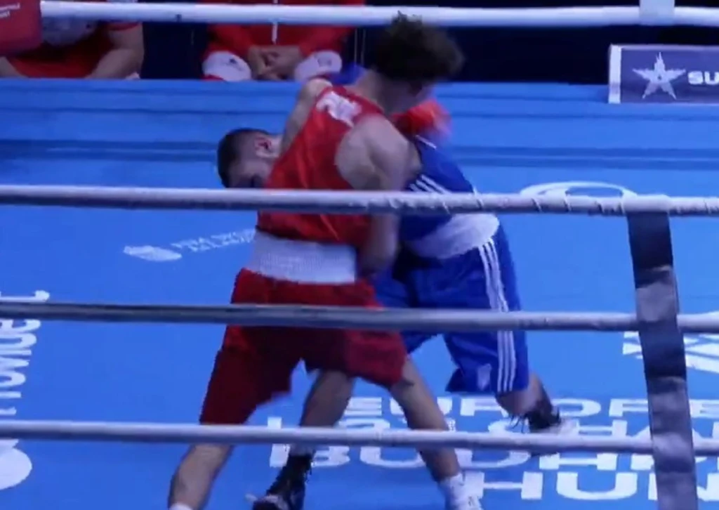 Dwóch bokserów w trakcie pojedynku na ringu; zawodnik w czerwonym stroju wykonuje cios w kierunku przeciwnika ubranego na niebiesko, wokół znajdują się liny ringu, na trybunach widoczna jest publiczność.