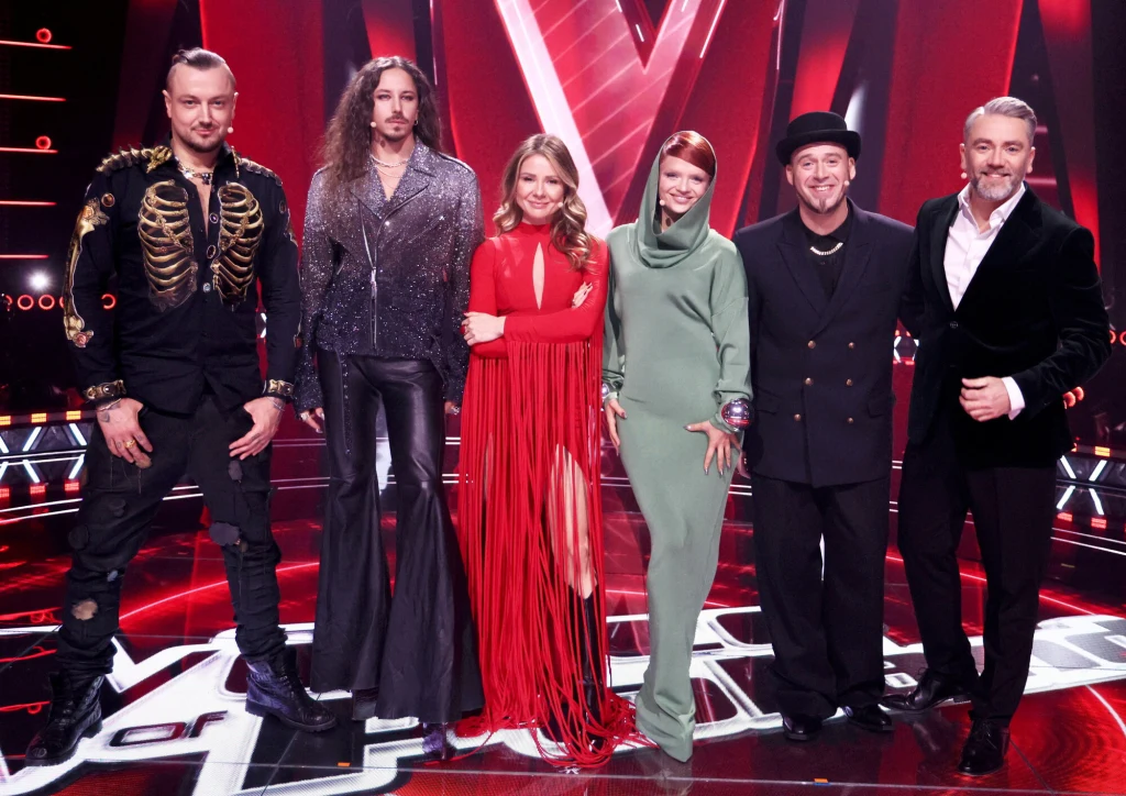 Trenerzy 16. edycji "The Voice of Poland": Baron, Michał Szpak, Ania Karwan, Margaret, Tomson i Kuba Badach Sześć osób ubranych w efektowne, sceniczne stroje stoi razem na scenie żywo oświetlonej czerwonym światłem, w tle zauważalne duże litery i dekoracje kojarzące się z programem muzycznym.