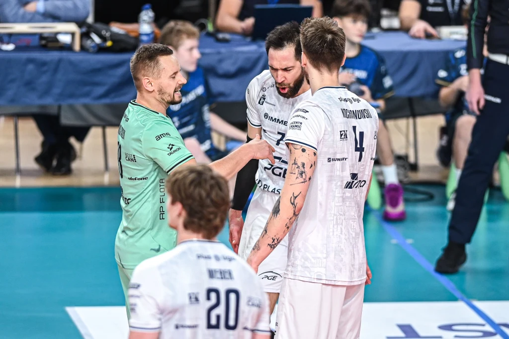 Projekt Warszawa Siatkarze drużyny w białych strojach oraz jeden zawodnik w zielonej koszulce stoją razem na boisku podczas rozmowy, w tle widoczni są członkowie sztabu oraz ławka rezerwowych.