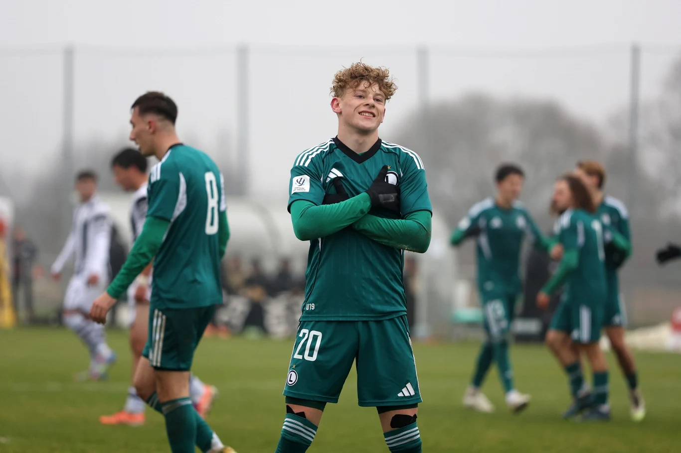 Młody piłkarz w zielonym stroju sportowym z numerem 20 stoi z założonymi rękami na boisku, wyrażając radość ze zdobycia gola, w tle pozostali zawodnicy w trakcie meczu piłkarskiego.