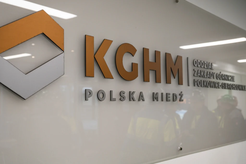Logo KGHM Polska Miedź z napisem Oddział Zakłady Górnicze Polkowice-Sieroszowice na jasnej ścianie, odbicia postaci w kaskach ochronnych widoczne w tle.