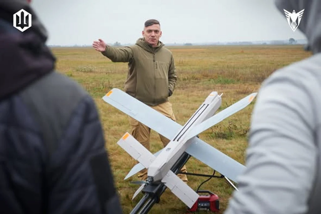 Mężczyzna w kurtce stoi na otwartym polu, gestykulując ręką, przed nim duży dron wojskowy na platformie startowej, w tle widać innych ludzi obserwujących sytuację.