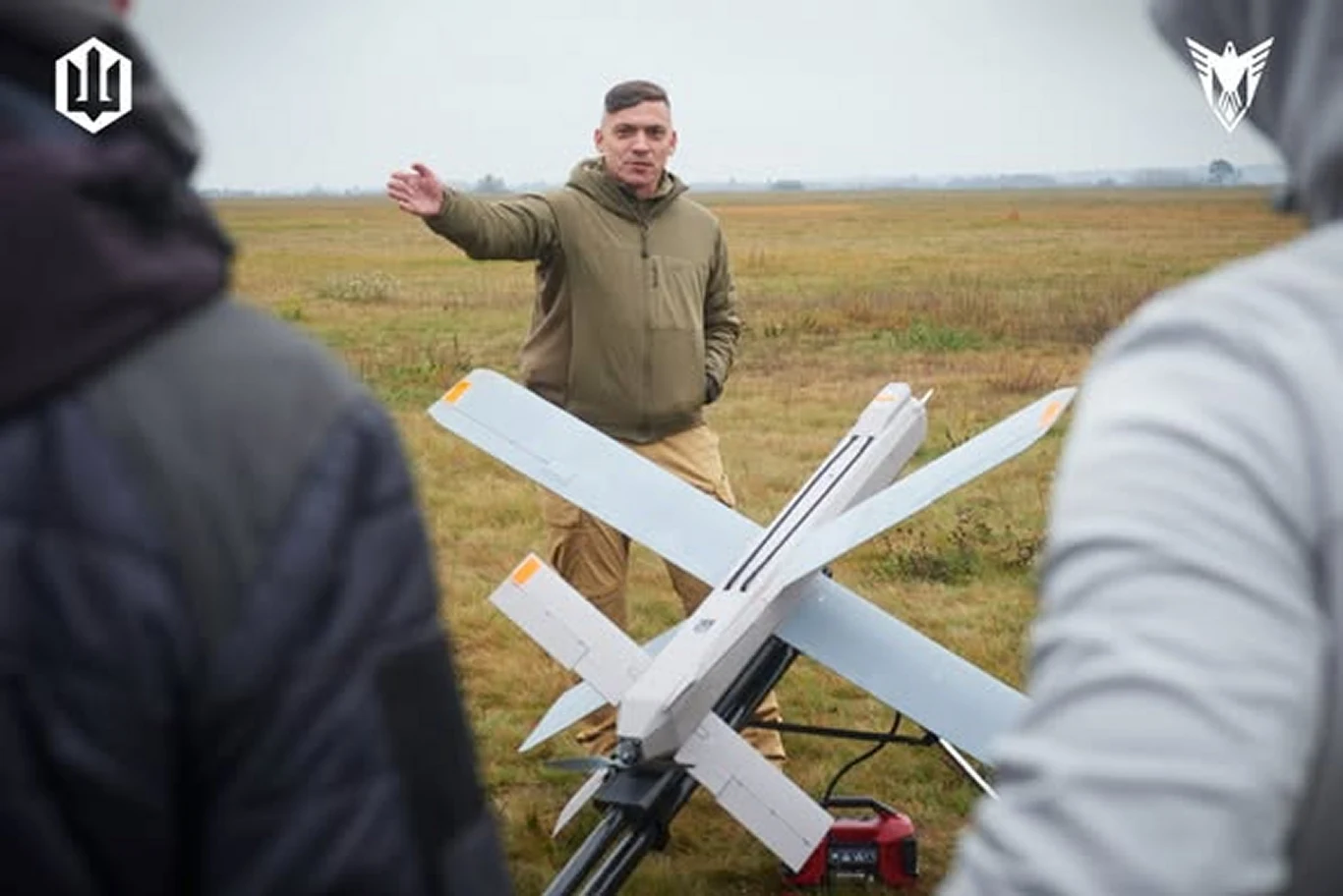 Skopiowali drona armii Putina i teraz atakują nim Rosję. Oto Shugai Mężczyzna w kurtce stoi na otwartym polu, gestykulując ręką, przed nim duży dron wojskowy na platformie startowej, w tle widać innych ludzi obserwujących sytuację.