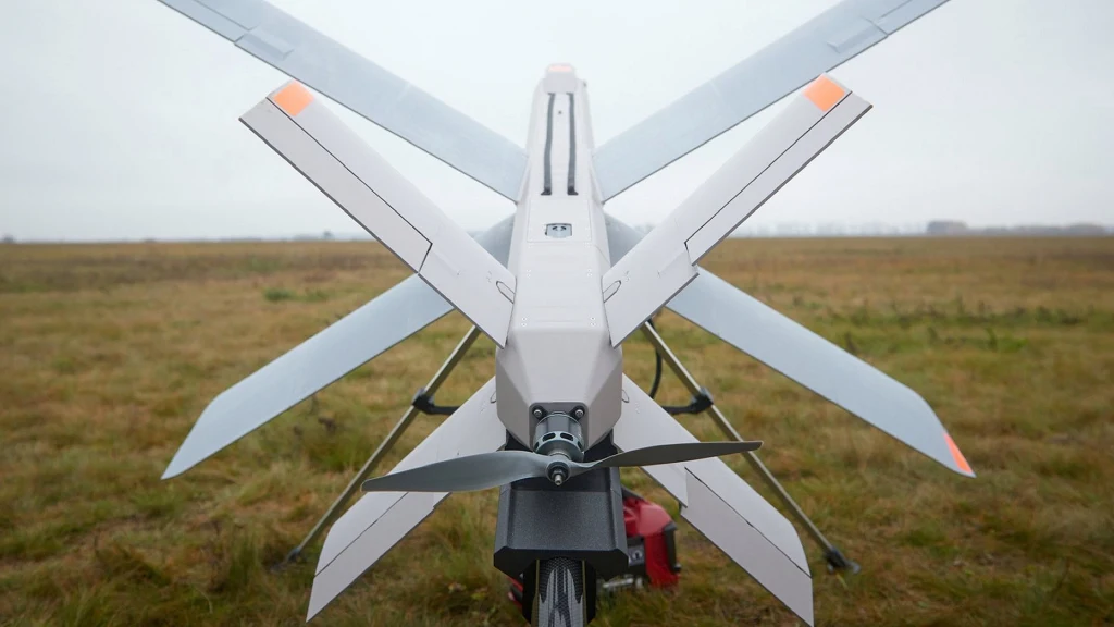 Bezpilotowy dron wojskowy ustawiony na trawiastym terenie, widok od tyłu z wyraźnie widocznymi śmigłami oraz rozłożonymi skrzydłami, w tle otwarta przestrzeń.