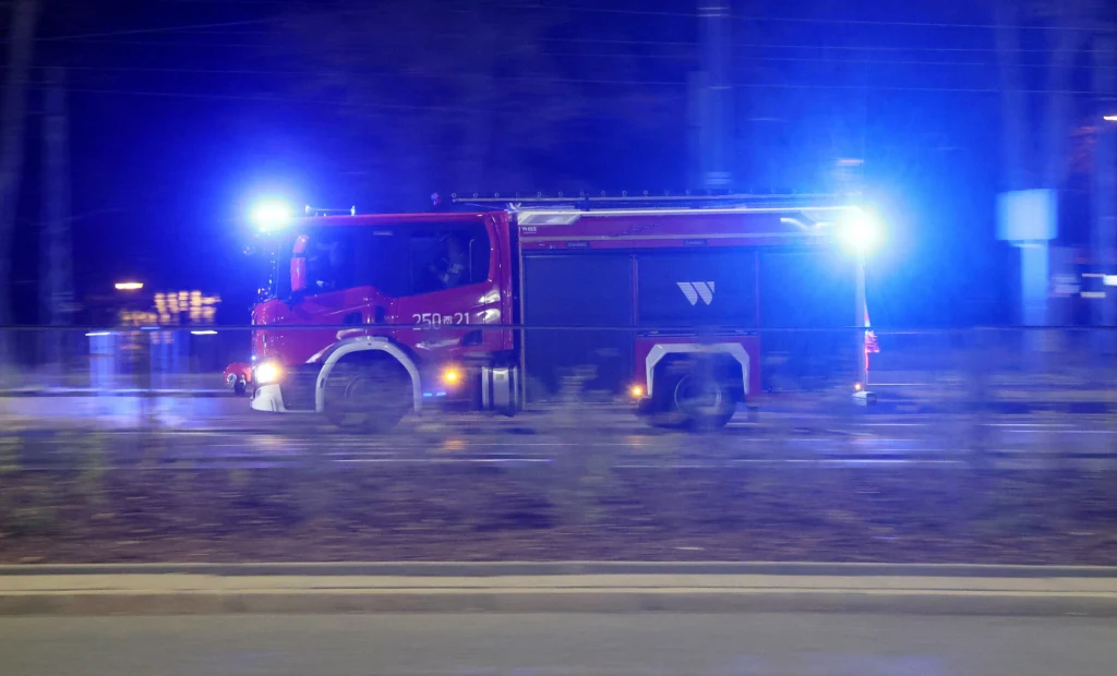 Czerwony wóz strażacki z włączonymi światłami sygnalizacyjnymi porusza się szybko po nocnej ulicy, światła niebieskie oświetlają okolicę, w tle widoczne ciemne drzewa i niewyraźne kontury miejskiego krajobrazu.