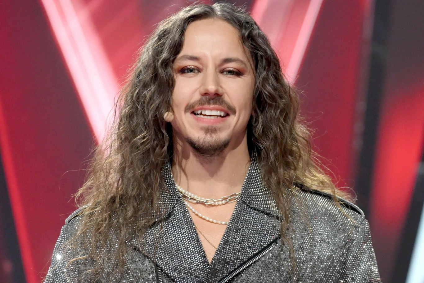 Michał Szpak świętuje swoje 35. urodziny Mężczyzna z długimi, falowanymi włosami, uśmiechnięty, ubrany w błyszczącą marynarkę z wieloma perłowymi naszyjnikami na szyi, widoczny na tle z czerwonymi i czarnymi akcentami graficznymi.