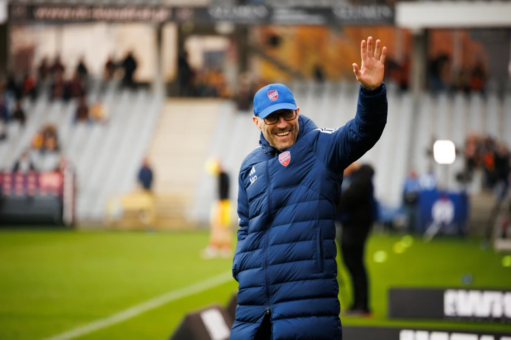 MArek Papszun chce odejsć do Legii Mężczyzna w długiej, pikowanej kurtce i czapce z daszkiem uśmiecha się i macha ręką na tle stadionu sportowego. W tle zamazane trybuny oraz niewyraźne postaci.