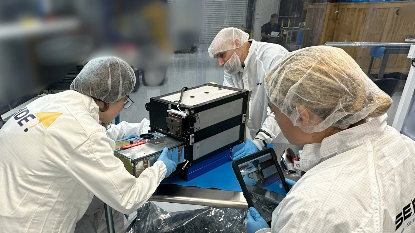 Satelita PW68 Trzy osoby w białych kombinezonach roboczych, maseczkach i czepkach wykonują prace montażowe lub badawcze przy zaawansowanym urządzeniu elektronicznym w laboratorium, korzystając z tabletu do dokumentacji.