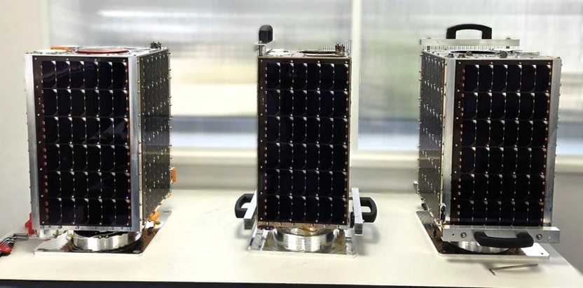 Satelity PIAST Trzy niewielkie satelity z panelami słonecznymi ustawione obok siebie na jasnym stole. Konstrukcje mają prostokątny kształt oraz widoczne uchwyty ułatwiające ich przenoszenie.