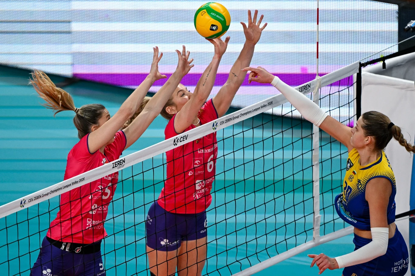 Kadr z meczu Ligi Mistrzyń siatkarek DevelopRes Rzeszów - Marica Płowdiw Trzy siatkarki w trakcie dynamicznej akcji przy siatce walczą o piłkę, dwie zawodniczki w czerwonych strojach próbują zablokować atakującej w żółto-niebieskim stroju.