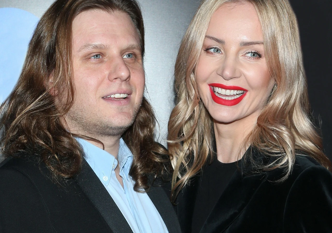 Dwoje dorosłych uśmiechniętych ludzi ubranych elegancko, kobieta z długimi blond włosami i wyrazistą czerwoną szminką, mężczyzna z dłuższymi brązowymi włosami, oboje pozują razem na jasnym tle.