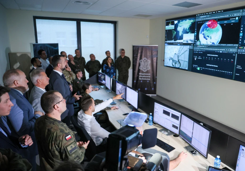 Grupa osób w mundurach i garniturach obserwuje duże monitory w nowoczesnej sali operacyjnej, na których wyświetlane są mapy, wykresy oraz wizualizacje danych, część osób pracuje przy komputerach, inni uważnie śledzą przebieg prezentacji.