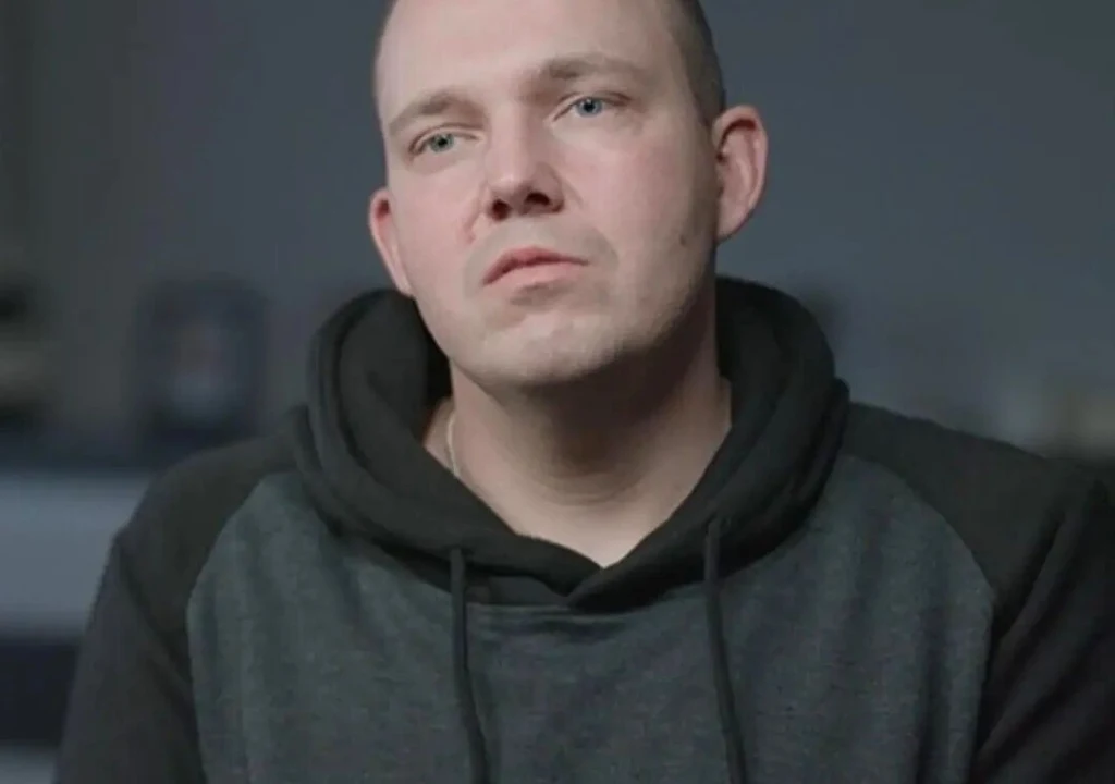 Krystian Plak, "Ślub od pierwszego wejrzenia" Mężczyzna w średnim wieku ubrany w ciemną bluzę z kapturem, patrzący przed siebie z neutralnym wyrazem twarzy, rozmyte tło w stonowanych kolorach.