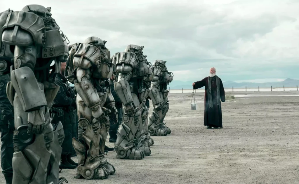 Kilku ludzi w zaawansowanych egzoszkieletach lub pancerzach stoi w szeregu na pustynnym terenie, naprzeciw mężczyzny ubranego w czarne szaty przypominające duchownego, który trzyma wiadro i unosi rękę w ich kierunku.
