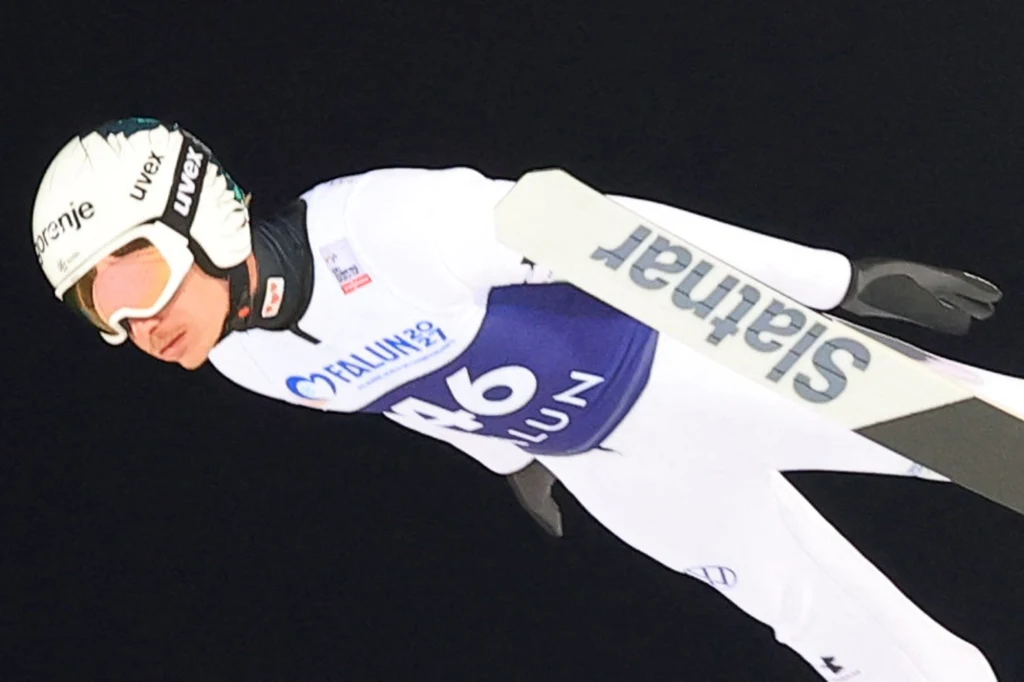 Anze Lanisek Zawodnik w białym kombinezonie i kasku podczas skoku narciarskiego w locie, na czarnym tle, widoczne narty oraz logotypy sponsorów na stroju i wyposażeniu sportowca.