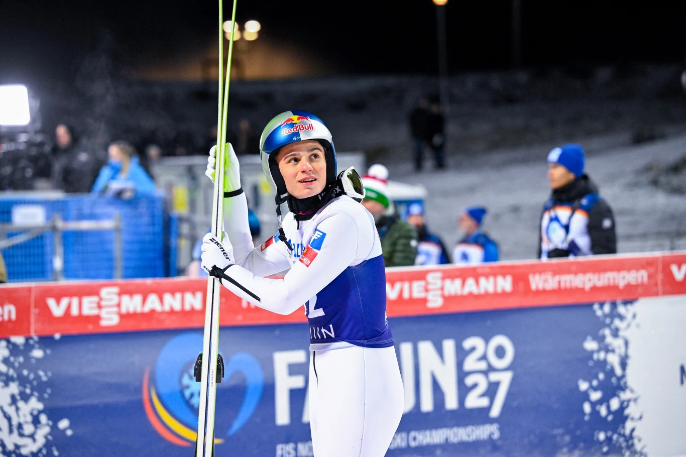Zawodnik w stroju narciarskim i kasku trzyma narty, w tle widoczny baner promujący mistrzostwa świata oraz osoby ubrana w odzież zimową, nocna sceneria z oświetleniem sztucznym.