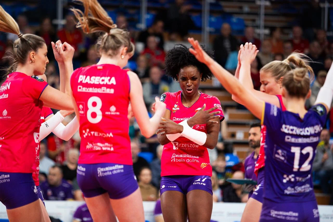 Siatkarki w strojach klubowych podczas meczu, skupione w kręgu z wyciągniętymi rękami, gotowe do zespołowego dopingowania lub uczczenia zdobytego punktu, w tle kibice na trybunach.