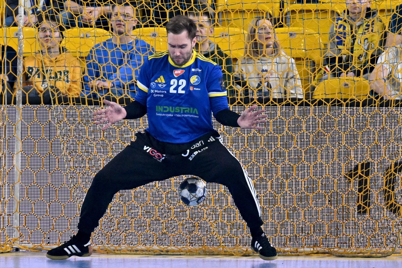 Bramkarz drużyny piłki ręcznej w niebieskiej koszulce z numerem 22 powstrzymuje piłkę przed bramką. Za nim widoczni kibice i żółte trybuny.