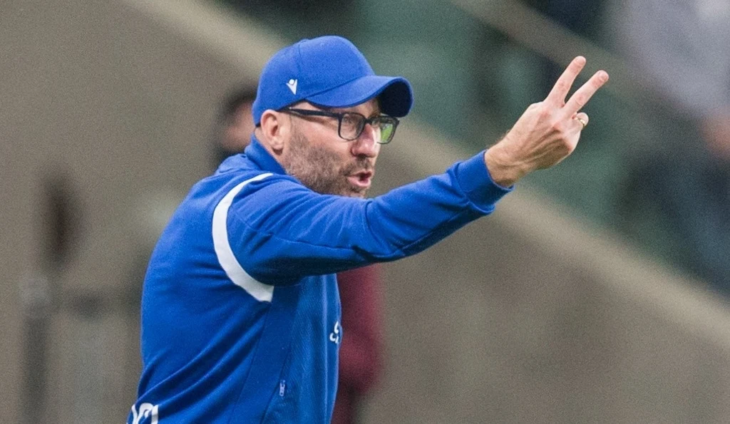 Marek Papszun Trener w niebieskiej sportowej kurtce i czapce gestykuluje ręką, pokazując dwa palce, patrzy w stronę boiska z poważnym wyrazem twarzy.