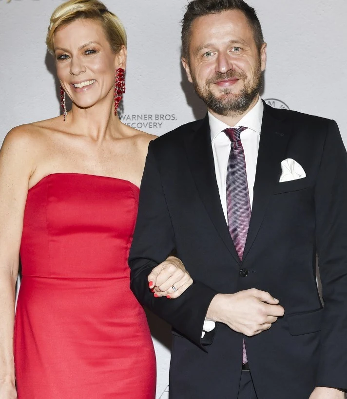 Anita Werner na baluTVN Elegancka kobieta w czerwonej sukni i mężczyzna w czarnym garniturze z białą poszetką oraz krawatem, stojący razem na tle ściany z logotypami, oboje uśmiechnięci i pozujący do zdjęcia.