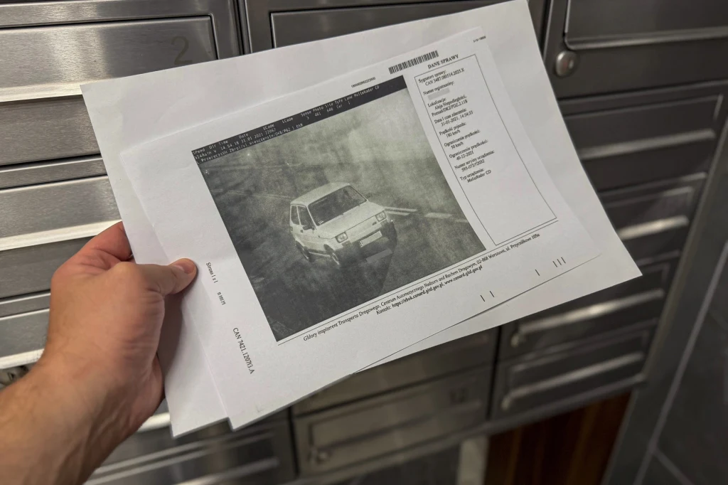 Ręka trzymająca wydrukowany na kartce fragment zdjęcia samochodu Fiat 126p oraz tabelę zawierającą szczegółowe informacje techniczne pojazdu, w tle widoczne metalowe skrzynki pocztowe.