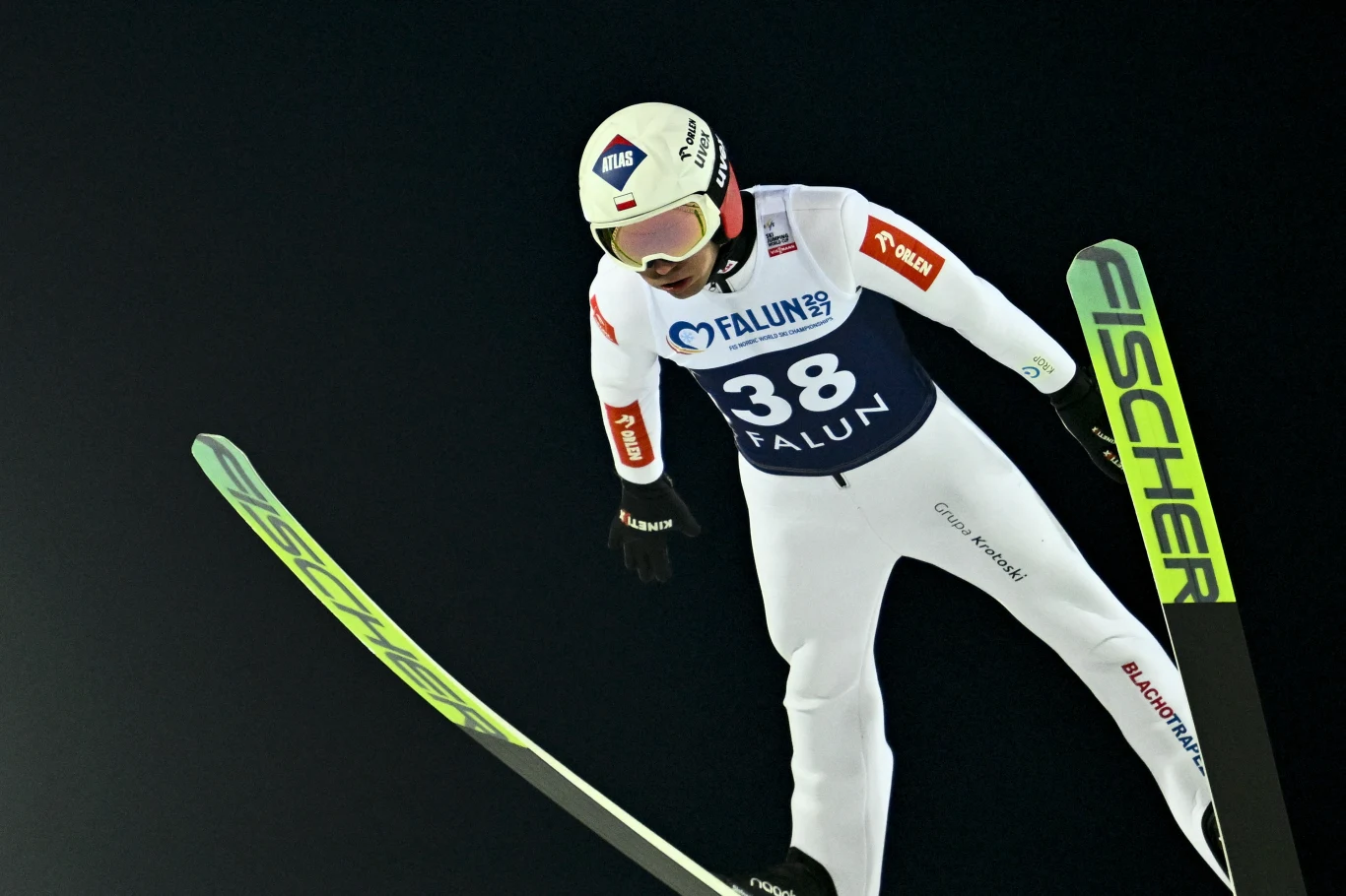 Kamil Stoch Zawodnik w białym kombinezonie sportowym leci w powietrzu na nartach podczas skoku narciarskiego, widoczny numer 38 oraz logotypy sponsorów na stroju.