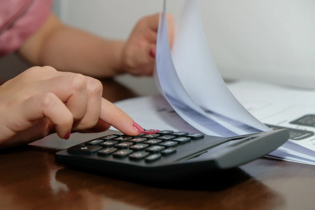 Dłoń osoby obsługującej kalkulator, obok której znajdują się dokumenty i kartki papieru, scena sugerująca pracę biurową lub rozliczenia finansowe.