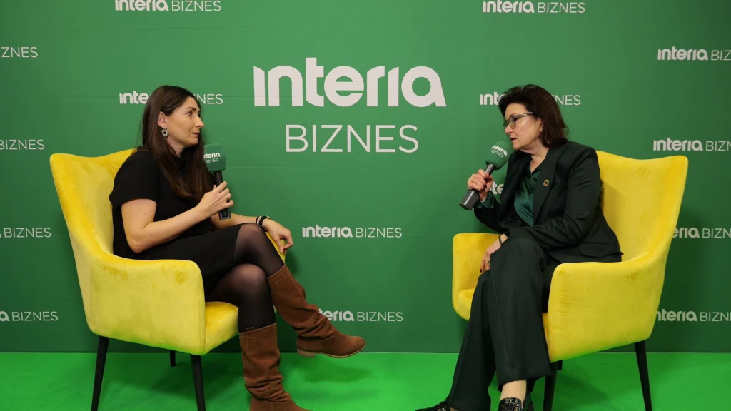Dwie kobiety siedzą na żółtych fotelach, prowadząc wywiad na tle zielonej ścianki z logo Interia Biznes. Obie trzymają mikrofony i są skierowane w swoją stronę, koncentrując się na rozmowie.