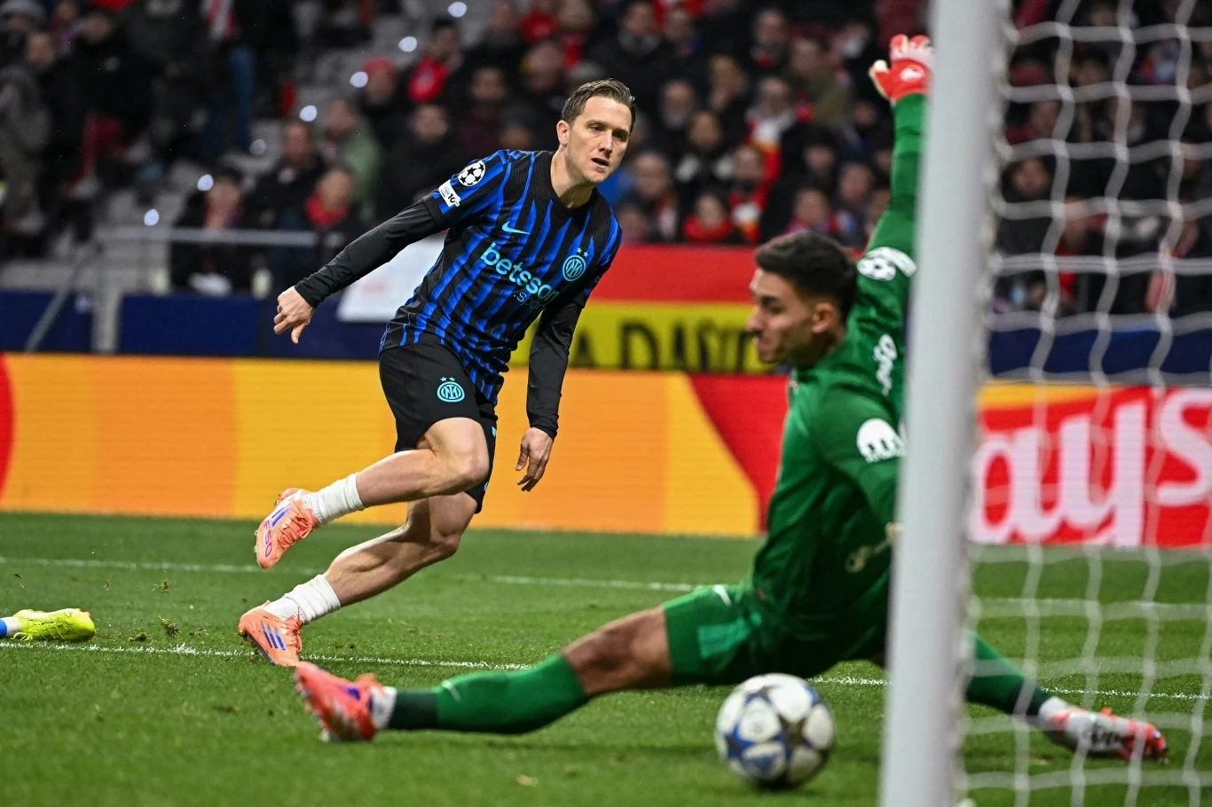 Piłkarz drużyny w czarno-niebieskich strojach oddaje strzał na bramkę, podczas gdy bramkarz w zielonym stroju próbuje obronić piłkę. W tle widać kibiców na trybunach oraz kolorowe reklamy przy boisku.