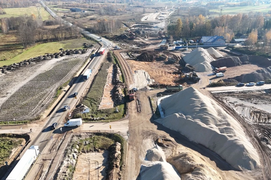 Rozległy plac budowy nowej drogi, widoczna infrastruktura drogowa oraz pojazdy budowlane i ciężarowe, wysokie hałdy piachu i żwiru, droga częściowo przebudowywana, otoczenie wiejskie z polami i lasami.