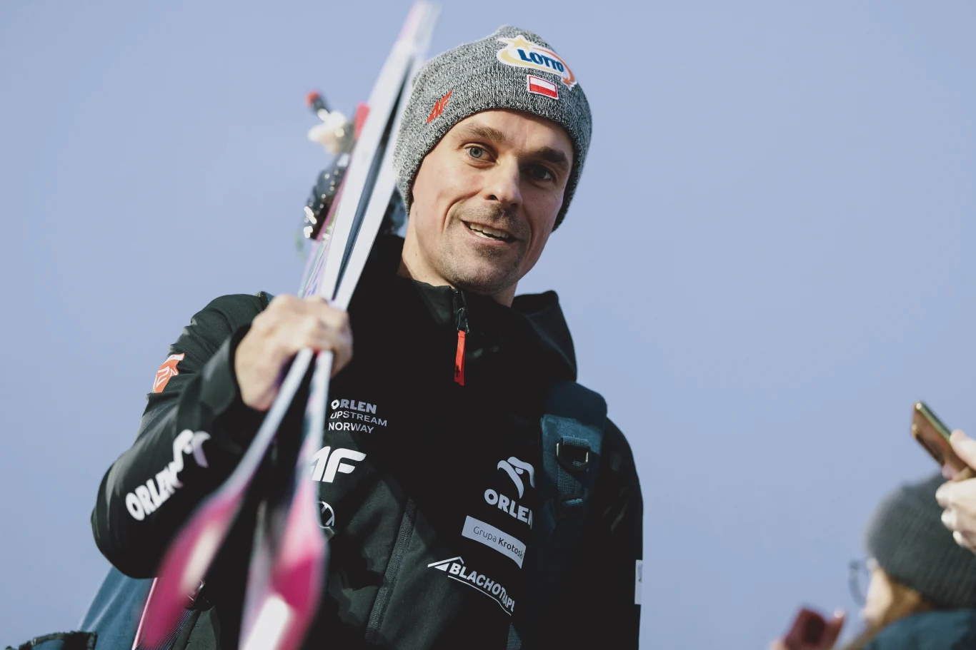 Piotr Żyła Mężczyzna w sportowej kurtce i czapce z logotypem, trzymający narty w ręku, w tle rozmyte sylwetki i niebo.