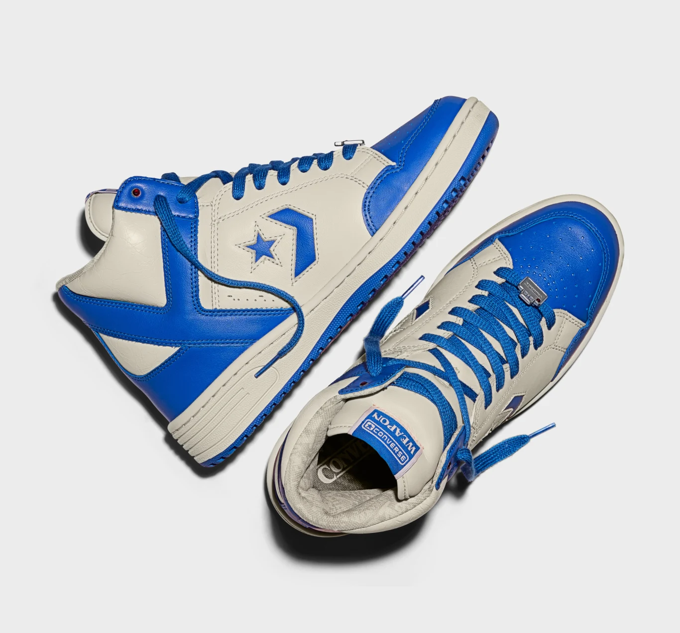 Converse Weapon x Stranger Things Para białych sportowych butów z niebieskimi elementami, widoczne logo Converse na boku oraz na języku, ustawione na jasnym tle jedna leżąca bokiem, druga od góry.