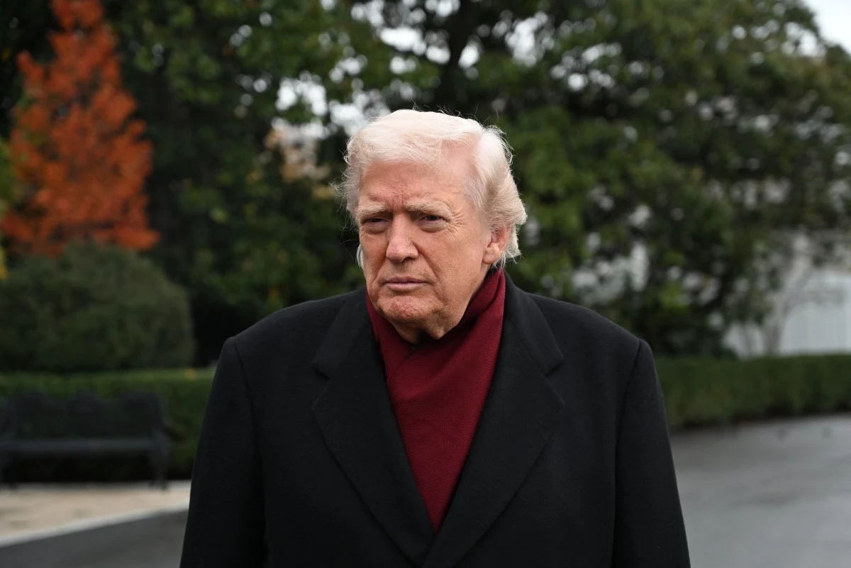 Prezydent USA Donald Trump Starszy mężczyzna ubrany w ciemny płaszcz i bordowy szalik stoi na zewnątrz, w tle drzewa i zieleń ogrodowa.
