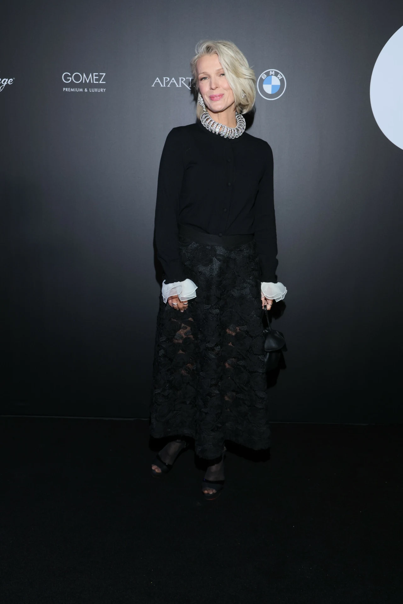 Magda Mołek w czarnym total looku. Patrzcie tylko na spódnicę Elegancko ubrana kobieta z jasnymi włosami w stylowej, czarnej spódnicy i czarnym swetrze ozdobionym dużym naszyjnikiem, stojąca na tle ściany z logotypami marek.