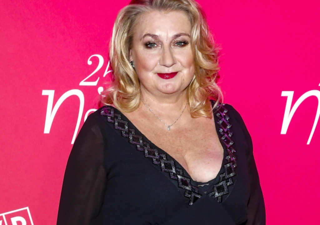 Kobieta w średnim wieku w eleganckiej czarnej sukni z dekoltem, na tle różowej ścianki z jasnym napisem.