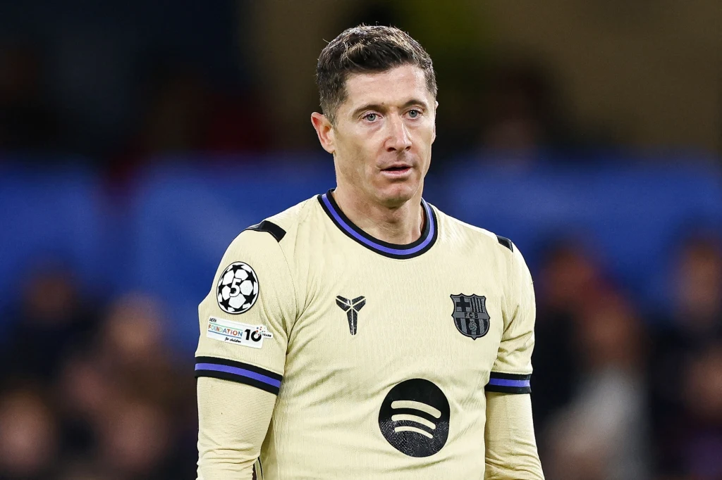 Piłkarz w jasnej koszulce drużyny FC Barcelona z widocznym logo sponsora oraz naszywkami na ramieniu, stoi na boisku na tle rozmytego stadionu.