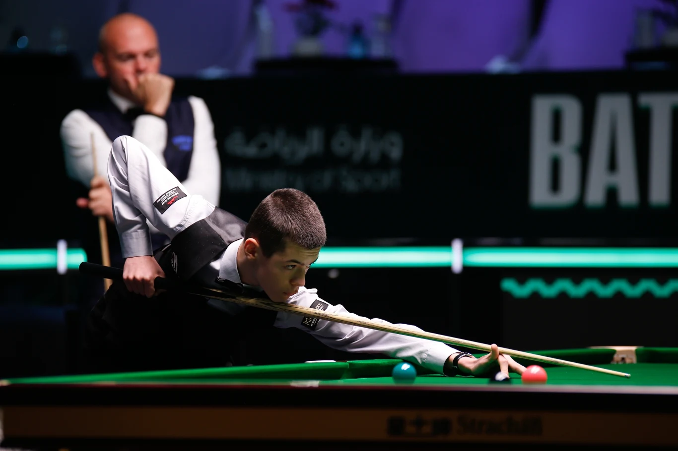 Młody snookerzysta wykonuje precyzyjne zagranie, skupiony nad stołem bilardowym, w tle sędzia obserwuje przebieg meczu. Kontrastowe oświetlenie podkreśla sportową atmosferę.