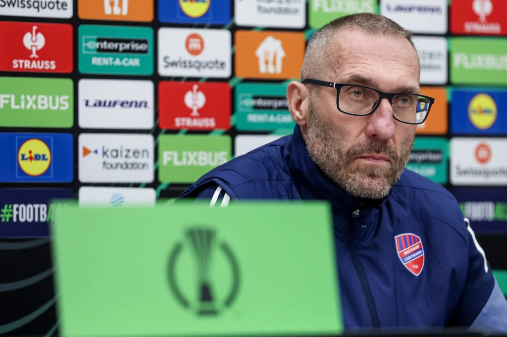 Marek Papszun przed meczem w Lidze Konferencji Mężczyzna w okularach i sportowej kurtce siedzi na tle tablicy z logotypami sponsorów oraz piłkarskimi akcentami, z przodu widoczny zielony element z logo turnieju piłkarskiego.