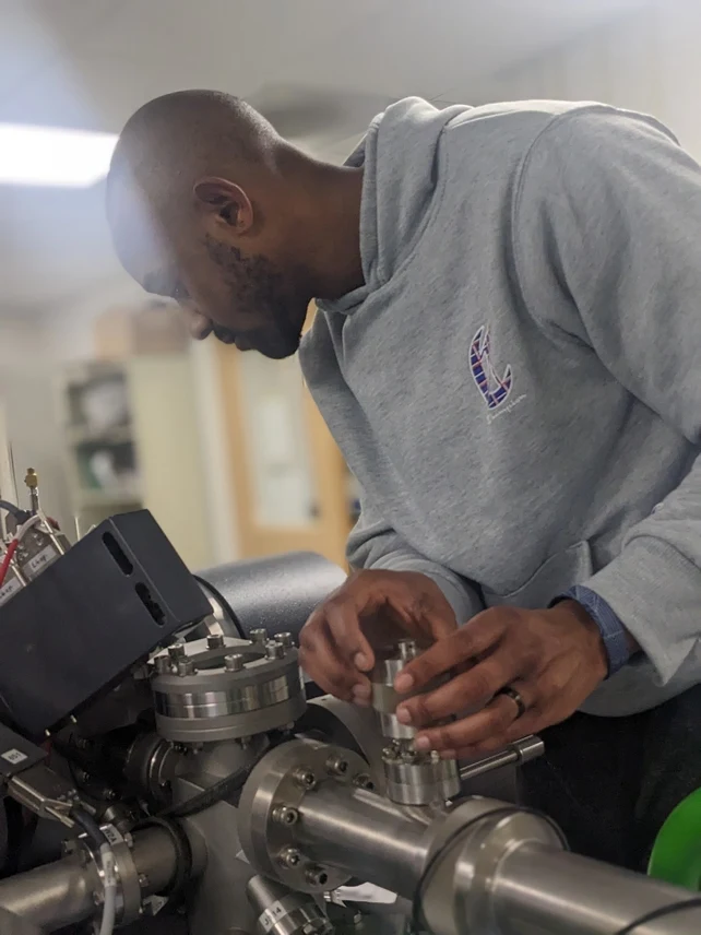 Mężczyzna w szarej bluzy pracuje przy skomplikowanej aparaturze laboratoryjnej, skupiając się na jej precyzyjnych elementach i ustawieniach.