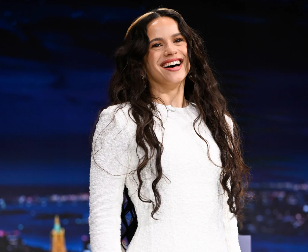 Rosalía promuje płytę "Lux" Uśmiechnięta kobieta o długich, falowanych, ciemnych włosach nosząca białą sukienkę. W tle widoczny rozmyty, kolorowy krajobraz nocnego miasta.