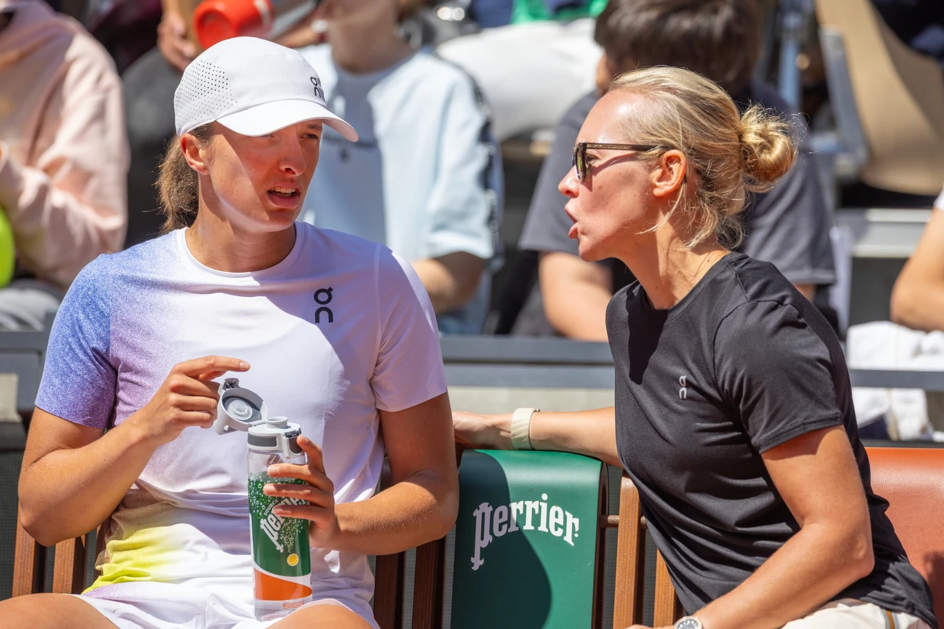 Iga Światek i Daria Abramowicz Dwie kobiety siedzą obok siebie na trybunach tenisowych, jedna w sportowym stroju z butelką w ręku, druga ubrana na czarno w okularach przeciwsłonecznych, rozmawiają ze sobą. W tle widoczni są kibice.