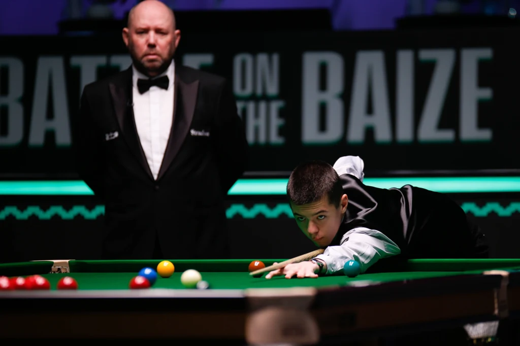 Zawodnik skupiony na wykonaniu uderzenia przy stole bilardowym podczas rozgrywek snookera, w tle sędzia w smokingu obserwuje sytuację na stole pod napisem 'Battle on the Baize'.