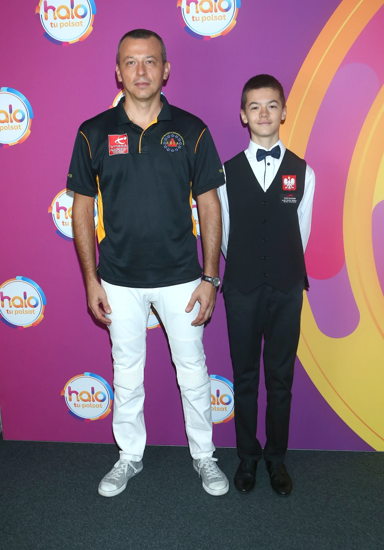 Dwie osoby stojące na tle kolorowej ścianki z logotypami, dorosły mężczyzna w czarnej koszulce sportowej i białych spodniach oraz nastolatek w czarnym garniturze z muchą i naszywką z orłem oraz polską flagą.
