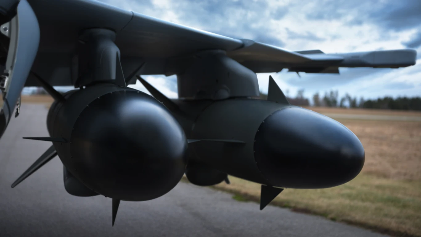 Podwieszone pod skrzydłem samolotu wojskowego bomby lub pociski z wyraźnie widocznymi aerodynamicznymi stabilizatorami, w tle rozmyty horyzont z trawiastym terenem i częściowo zachmurzonym niebem.