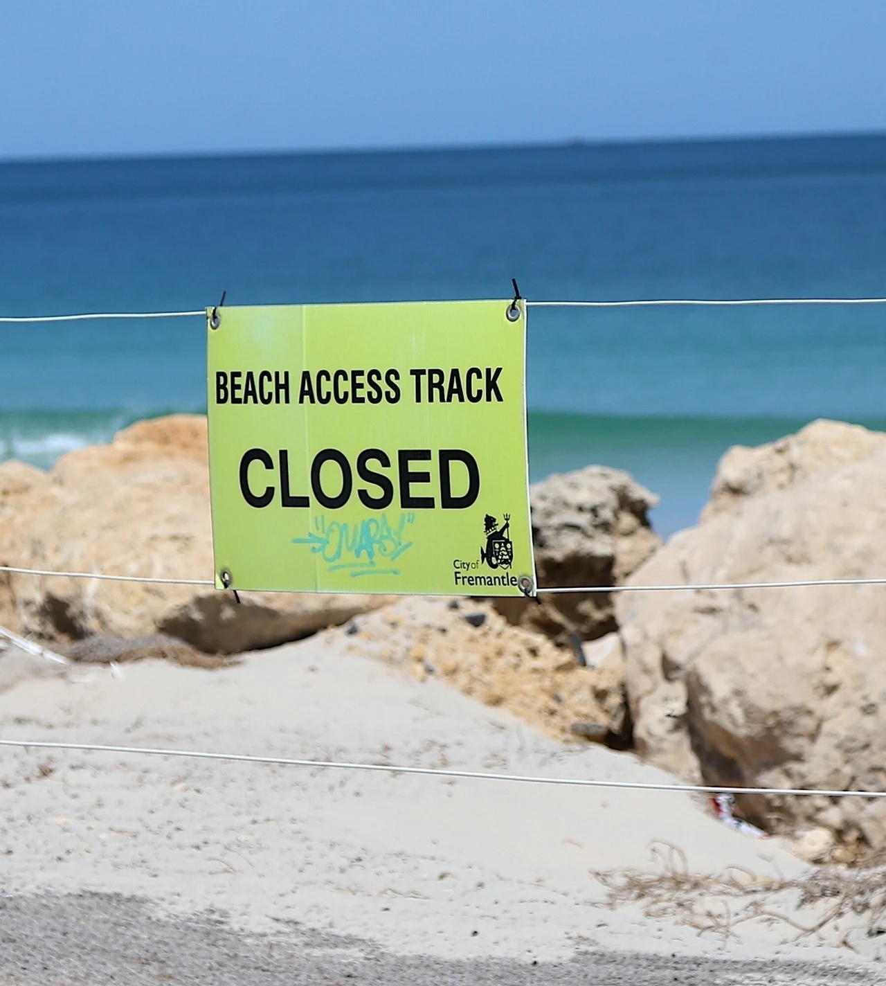 Władze natychmiast zamknęły plażę Nadmorska ścieżka prowadząca na plażę zamknięta z powodu wywieszonego znaku informacyjnego z napisem „Beach Access Track Closed”; w tle widoczna linia brzegowa, fale oraz duże skały.
