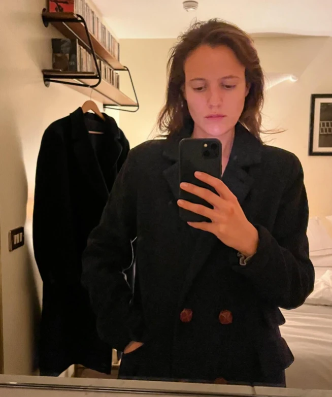 Osoba w ciemnym płaszczu robi selfie w lustrze, stojąc w przytulnie urządzonym pokoju z łóżkiem, w tle widoczny drugi płaszcz na wieszaku oraz półka z książkami lub płytami.