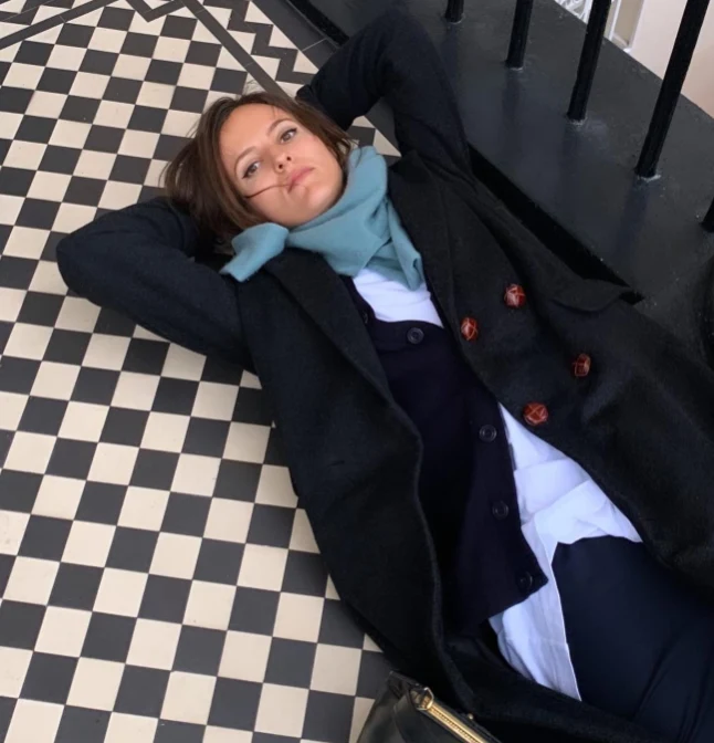 Kobieta ubrana w ciemny płaszcz i szalik leży na biało-czarnej, geometrycznej posadzce z rękami założonymi za głowę, obok widoczne są metalowe barierki.