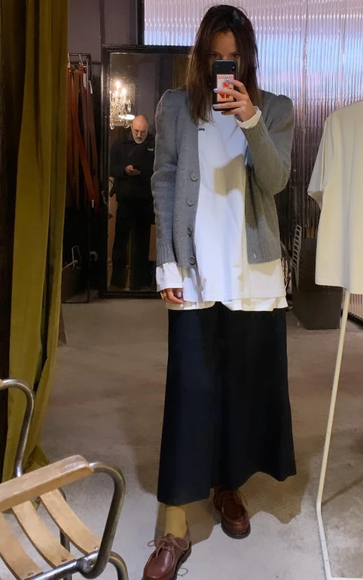 Młoda kobieta wykonuje zdjęcie selfie w lustrze, ubrana w szary kardigan, białą koszulę, czarną długą spódnicę i brązowe buty, w tle widać starszego mężczyznę patrzącego w stronę telefonu oraz elementy wnętrza sklepu odzieżowego.