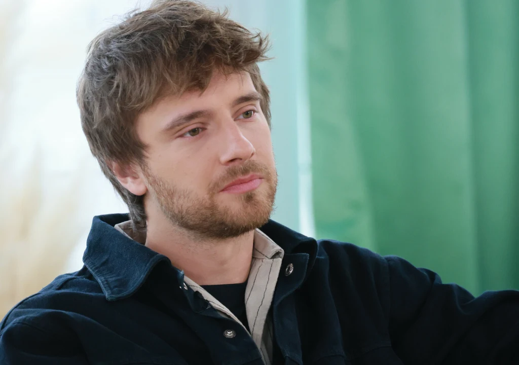 Młody mężczyzna z brodą i nieco rozczochraną fryzurą siedzi w pomieszczeniu ubrany w ciemną kurtkę i jasną koszulę, na tle zielonej zasłony.